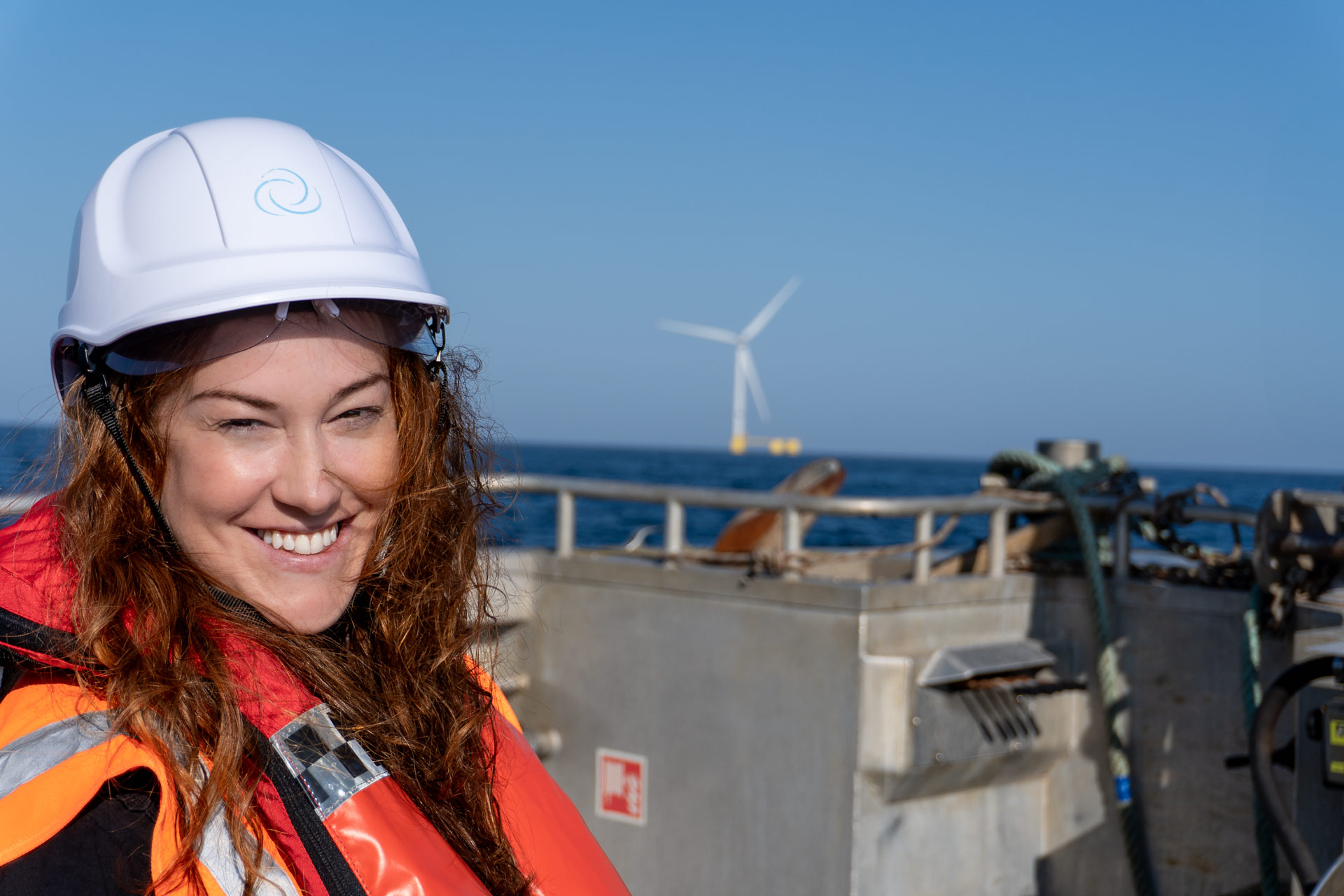 Flotation Energy team member in safety gear smiling on a vessel with a floating offshore wind turbine in the background